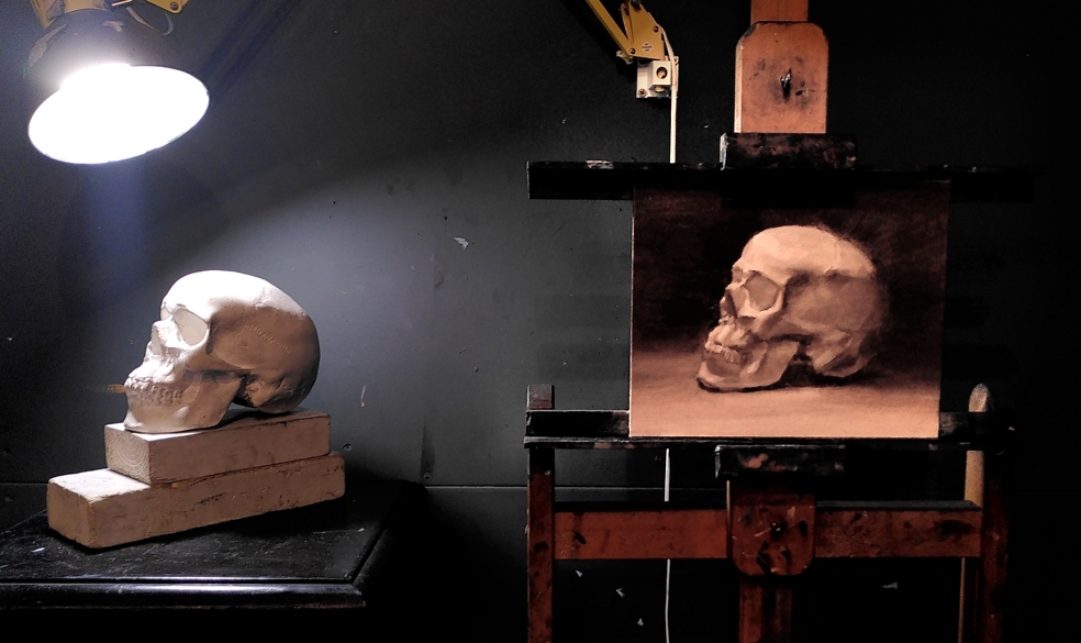 Grey-scale oil painting of a lit plaster cast human skull, beside the real thing.