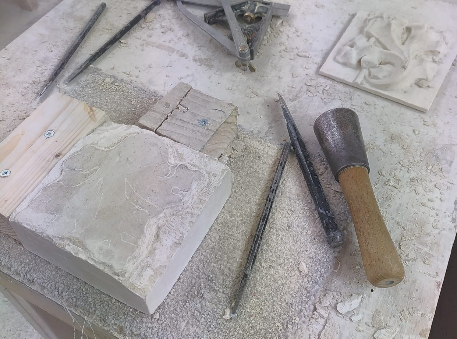 A square lump of Portland stone, a mallet, some chisels, and a relief tile with a gothic oak leaf design, sit on a dusty workbench.