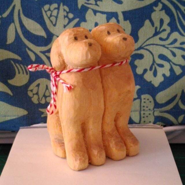 A whittled basswood statue of two identical dogs, standing side-by-side. They have pencil dots for eyes and noses, and are sharing a red and white thread scarf.