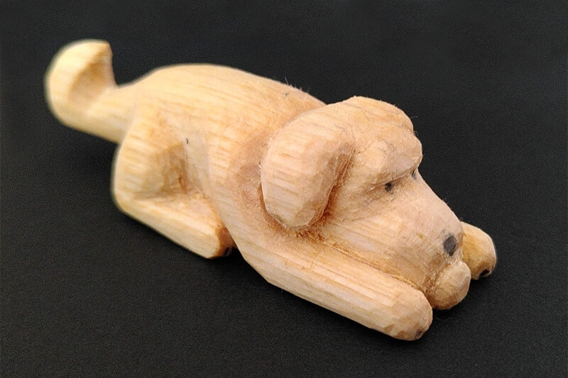 A whittled basswood statue of a playful prone dog with a ball, tail raised.