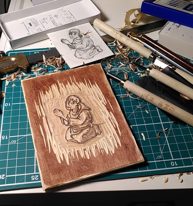 An in-progress woodcut of a kneeling monk. Carving tools and shavings surround the stained wood panel. A pencilled design lays beside the work area.