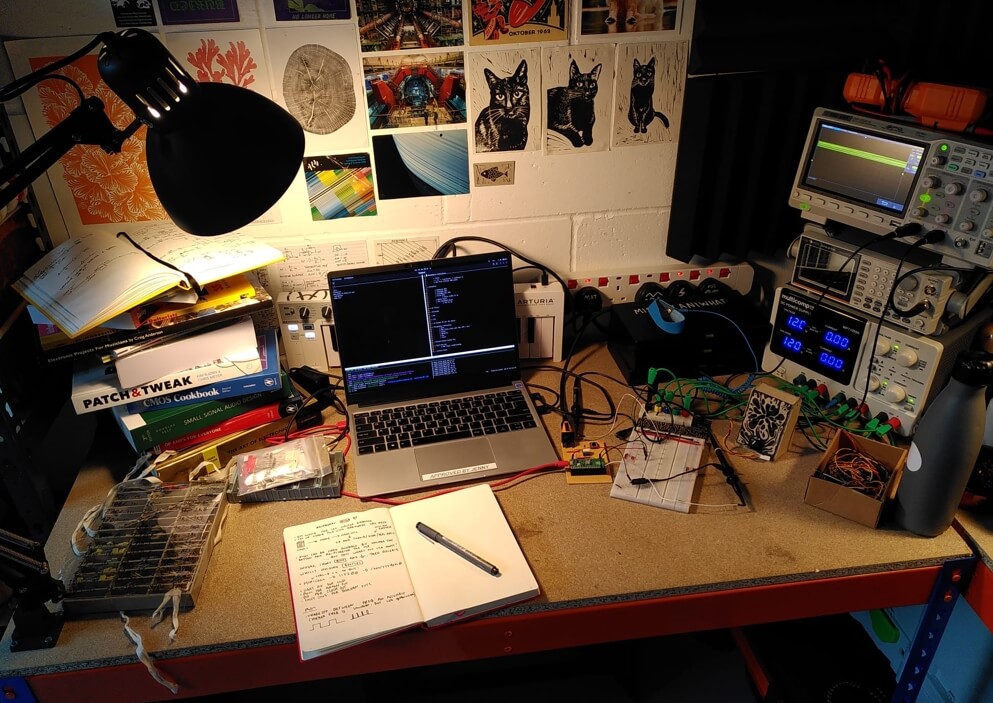 My desk, covered in electronics components, below some colourful posters of cats and scientific equipment.